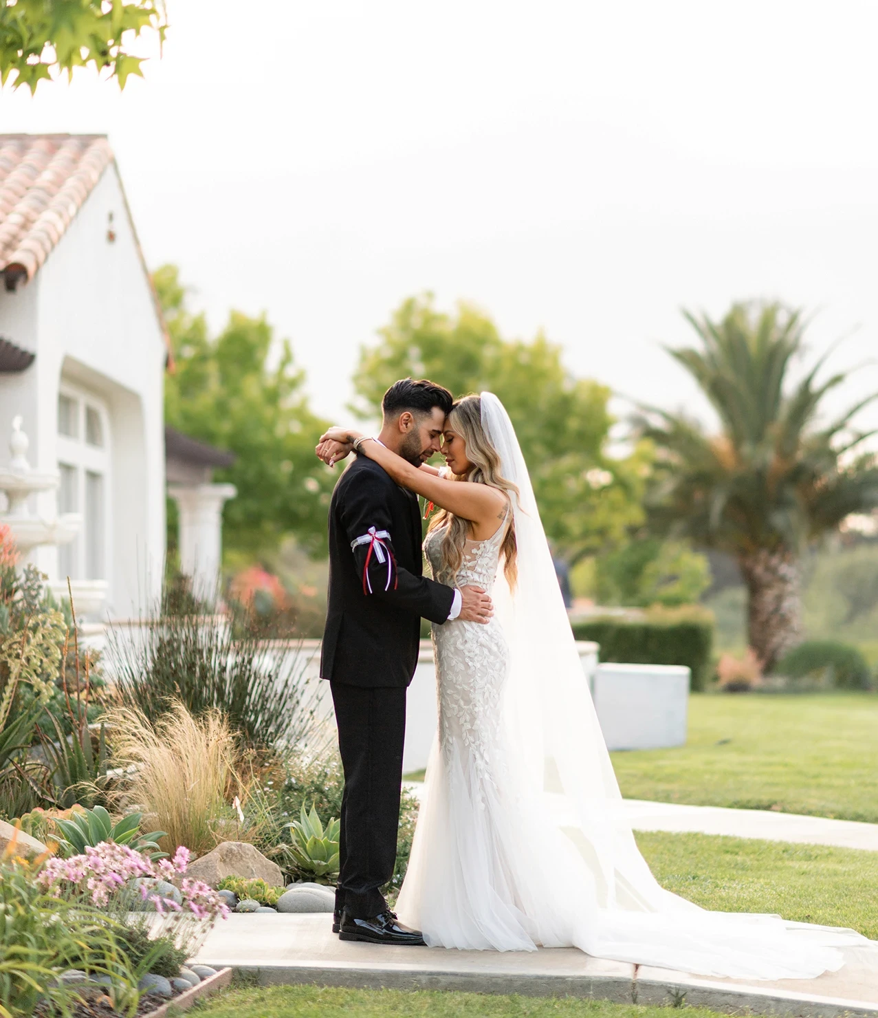 Bride and groom embracing outdoors on a pathway next to a white stucco building.