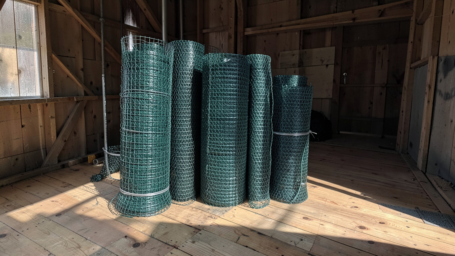 Rolls of green vinyl-coated crab pot wire mesh and galvanized rigging wire on cedar planks in a coastal gear shed.