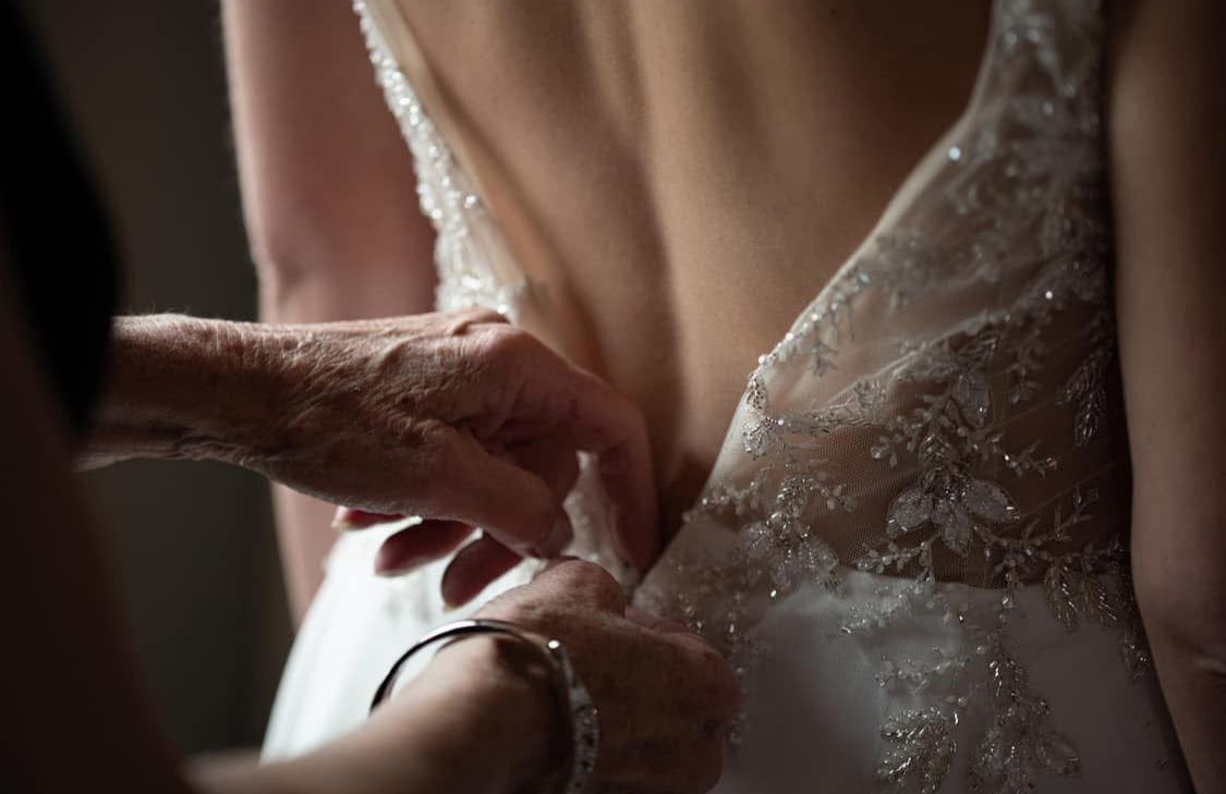 Hands helping a bride fasten the back of her beaded wedding dress.