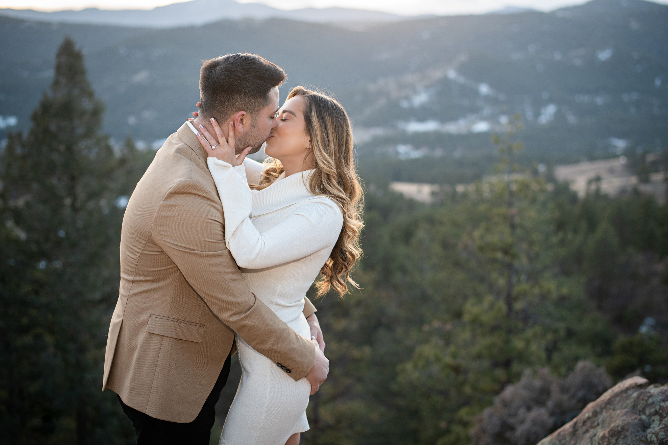 An engaged couple kissing outdoors with a scenic mountain backdrop during an engagement session.