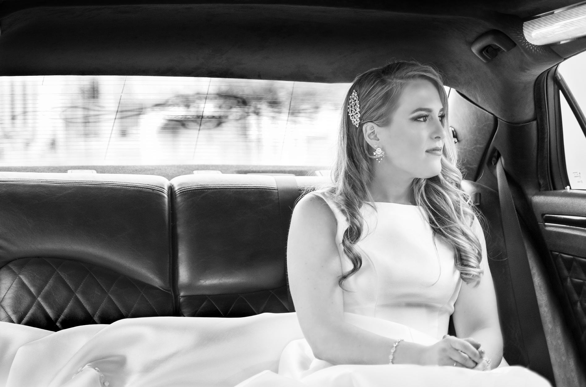 Bride in a white gown sitting in the back of a car looking out the window.