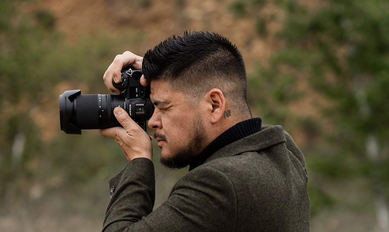 Photographer holding a Nikon camera to his eye while outdoors in a green blazer.