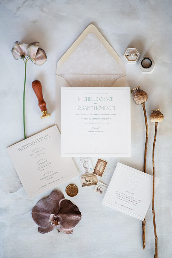 Flat lay of luxury wedding stationery, envelope, rings, and dried florals.