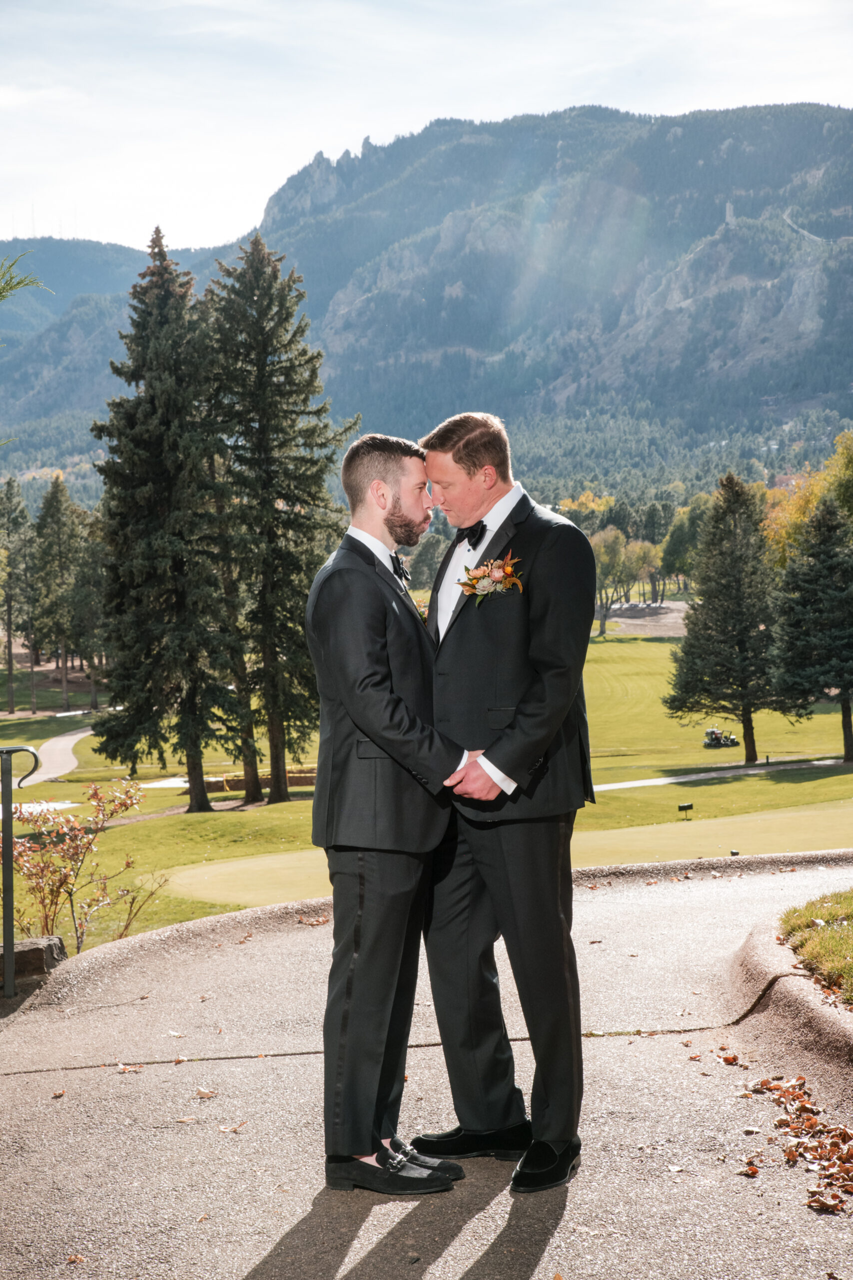 Two grooms in black tuxedos stand forehead to forehead on an outdoor path with mountain views.