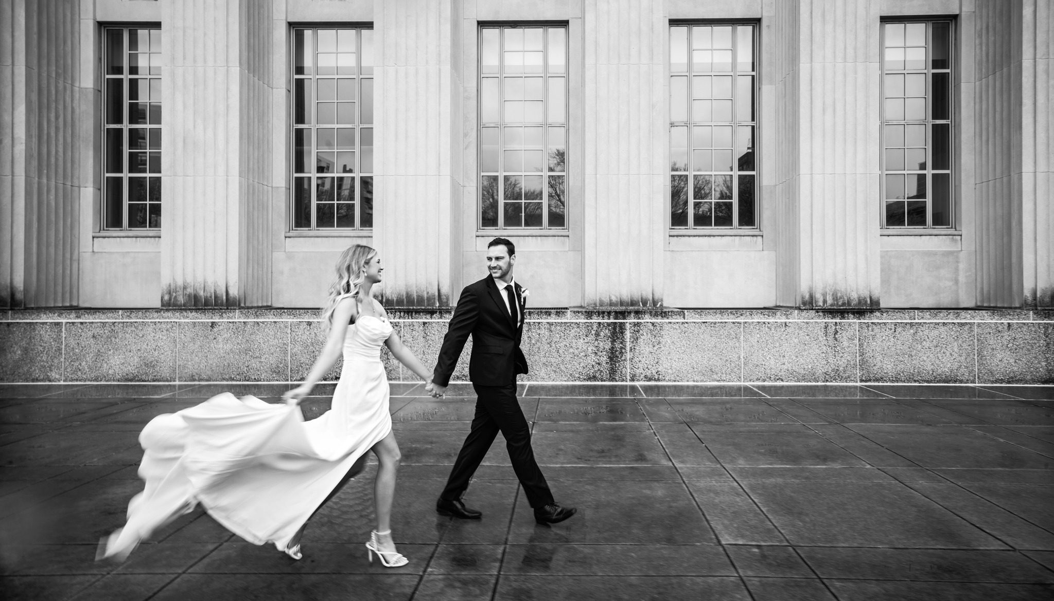 Bride and groom walking hand-in-hand outside a classic stone building.