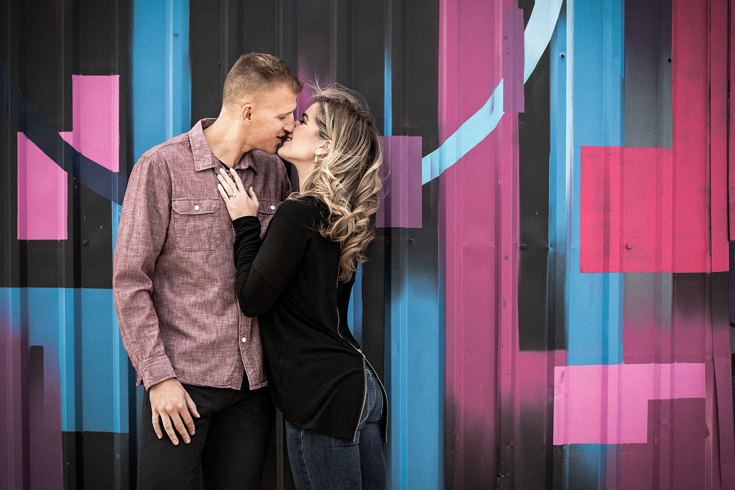 Engaged couple kissing in front of a colorful painted mural.